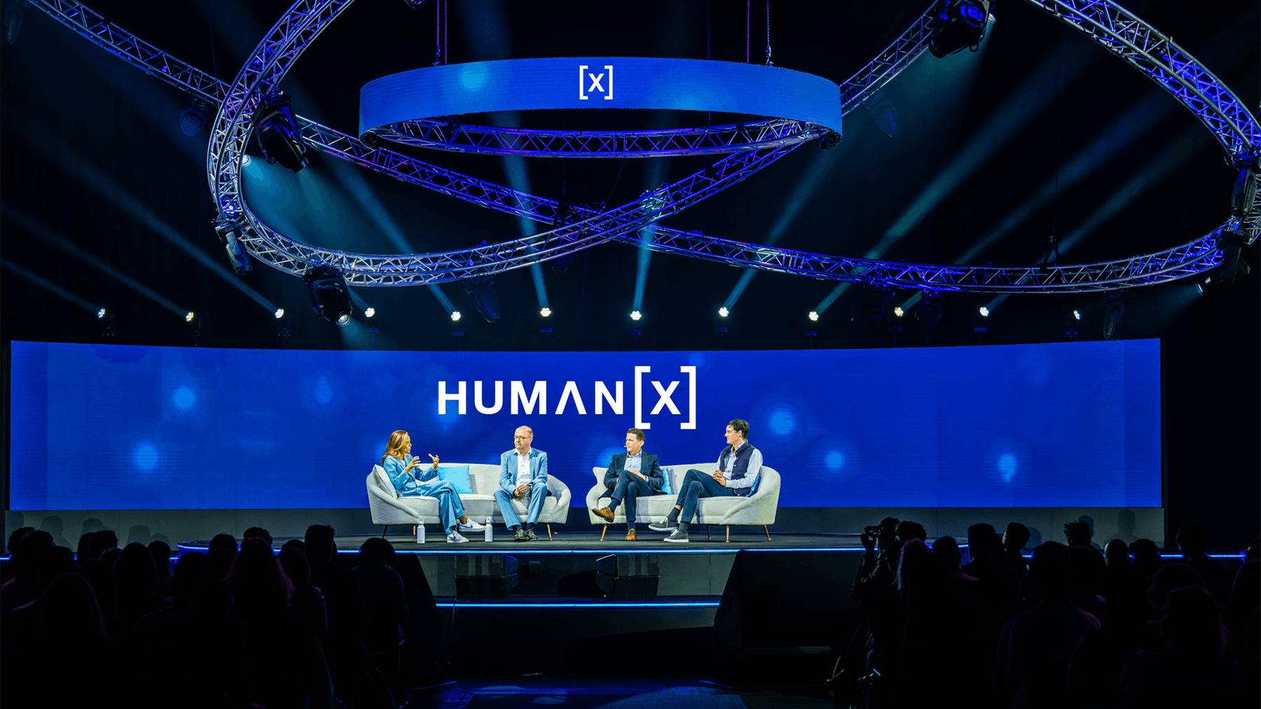 A panel of four speakers is seated on a stage with blue lighting and a large screen behind them displaying the conference name, "HUMAN [X]." The setting includes modern seating and overhead circular truss lighting. The audience is visible in the foreground.